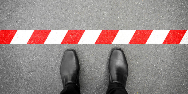 Se ven los zapatos de un hombre trajeado frente a una línea roja y blanca, como si no pudiese cruzarla. Está parado y de pie.